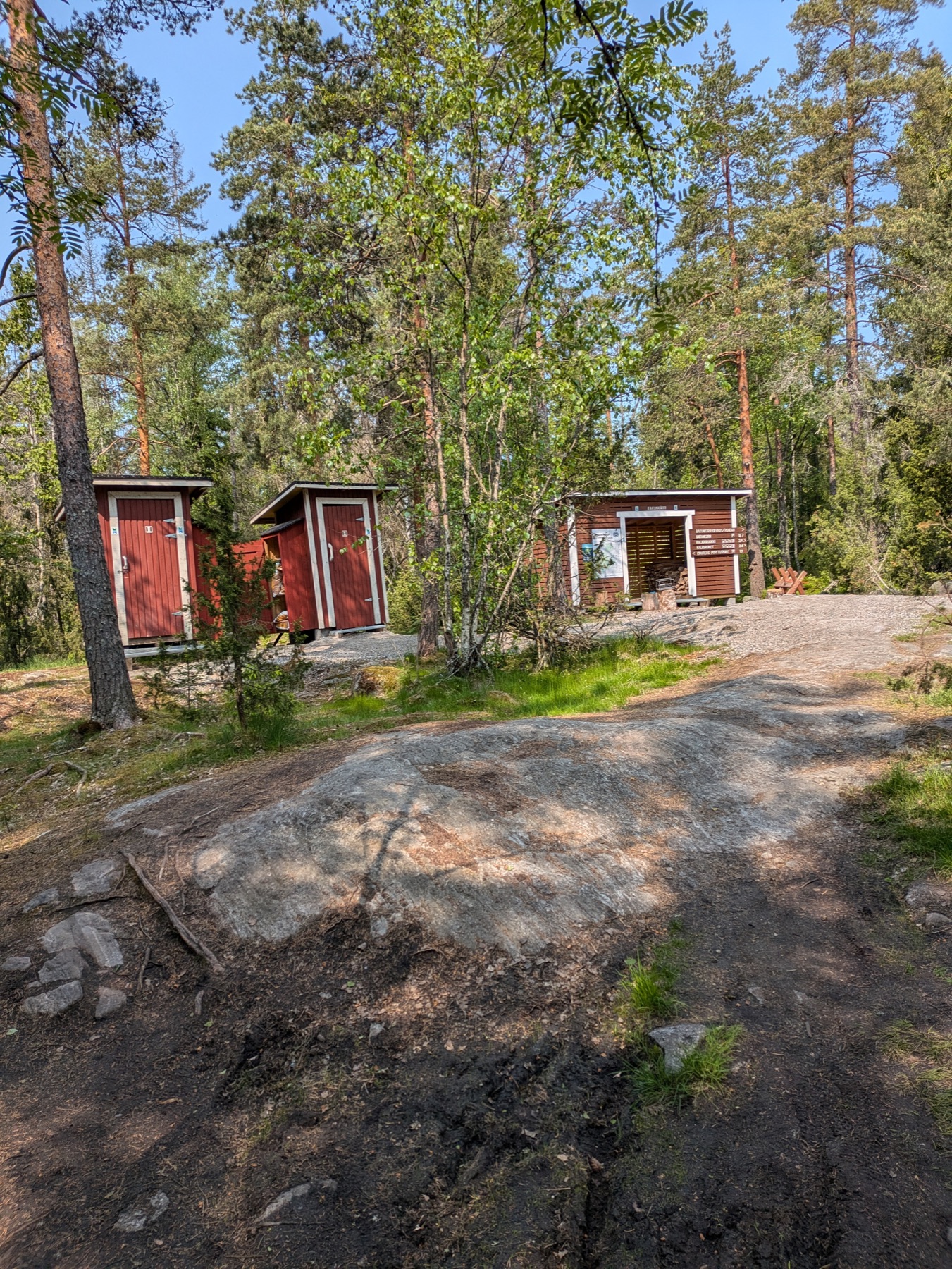 A forest campsite with red wooden huts and a firewood shelter on a sunny day